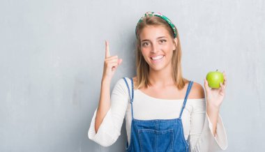 Güzel genç kadın grunge yüzünden yeşil elma bir fikirle sürpriz yemek duvar gri veya gülen yüz, bir numara ile işaret parmağı soru