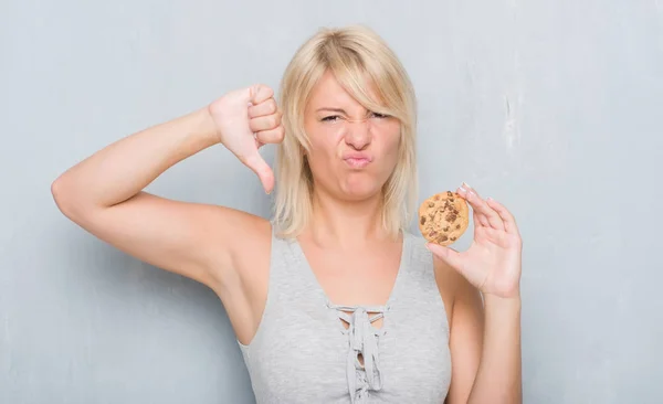 灰色グランジ以上白人の大人女性壁の怒った顔 ダウン親指で嫌悪感を示すマイナス記号 拒絶反応の概念とチョコレート クッキーを食べる — ストック写真