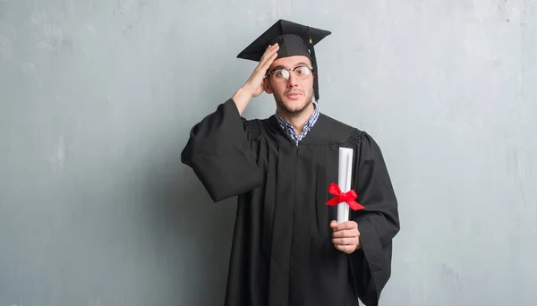 頭の上の手でストレス度を保持している大学院の制服を着て灰色グランジ壁を越えて若い白人男が恥と驚きの顔でショックを怒っているとイライラします 恐怖と動揺の間違い — ストック写真