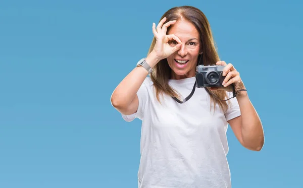 指の間から見て目に手で サインをして笑っている幸せそうな顔で孤立した背景にビンテージ写真カメラを使用して写真を撮る中年ヒスパニック系女性 — ストック写真