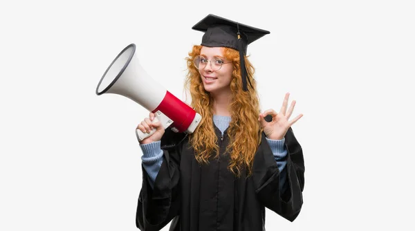 身に着けている赤毛の若い学生女性卒業制服持株メガホン優秀なシンボルの指で サインをしています — ストック写真