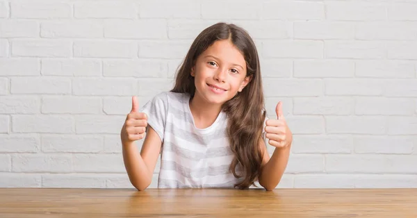 Young hispanic kid sitting on the table at home success sign doing positive gesture with hand, thumbs up smiling and happy. Looking at the camera with cheerful expression, winner gesture.