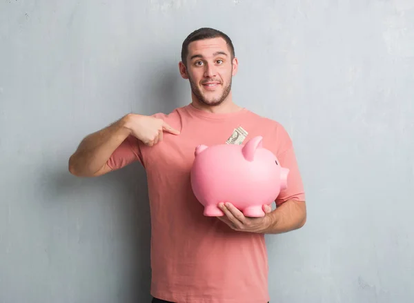 Joven Caucásico Hombre Sobre Gris Grunge Pared Holding Piggy Banco —  Fotos de Stock