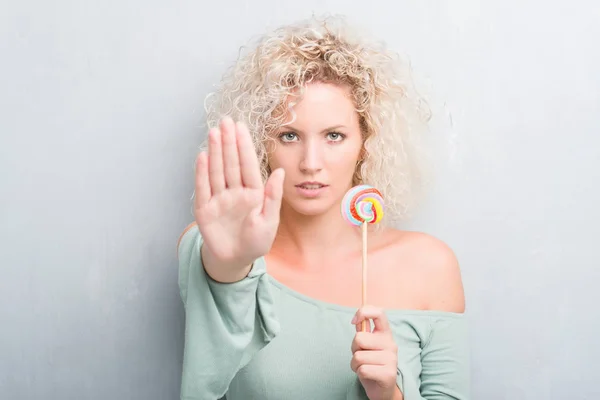 年轻的金发女子超过垃圾灰墙吃糖果棒糖与张开手做停止标志与严肃和自信的表达 防御手势 — 图库照片