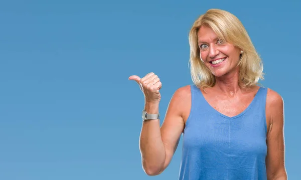 Middle age blonde woman over isolated background smiling with happy face looking and pointing to the side with thumb up.