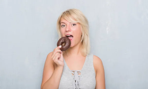 Caucasian adult woman over grey grunge wall eating chocolate donut with a confident expression on smart face thinking serious