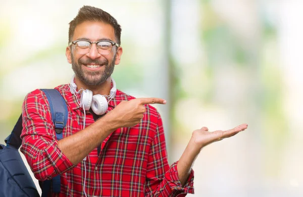 Hayret ve el ile sunulması ve parmak ile işaret ederken kameraya gülümseyen izole arka plan üzerinde kulaklık ve sırt çantası giyiyor yetişkin İspanyol öğrenci adam.