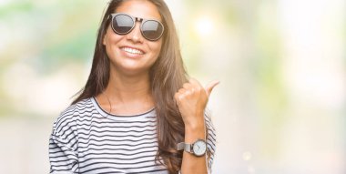 Genç güzel Arap kadın arıyorum ve başparmak yanında yukarıyı mutlu yüz ile gülümseyen izole arka plan üzerinde güneş gözlüğü takıyor.