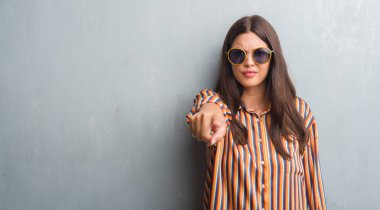 Grunge gri duvar retro güneş gözlüğü ve kamera ve size parmak ile işaret kıyafet giyen genç esmer kadın, el işareti, pozitif ve kendinden emin bir jest ön