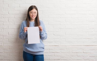 Genç Çinli kadın ayakta ve diş gösterilen kendine güvenen bir gülümsemeyle gülümseyerek mutlu bir yüz ile boş kağıt tutan tuğla duvarın üzerinden