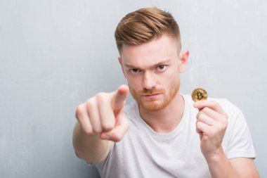 Genç Kızıl saçlı adam kamera ve size, el işareti, pozitif ve kendinden emin bir jest ön parmak ile işaret bitcoin gösterilen gri grunge duvarın üzerinden