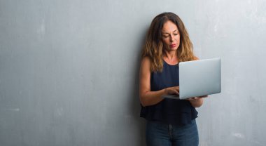 Acımak yüz ciddi düşünüyorum üstünde kendine güvenen bir ifade ile dizüstü kullanarak gri grunge duvar üzerinde duran orta yaş İspanyol kadın