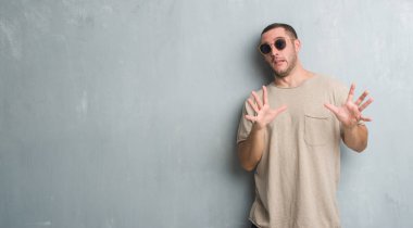 Şokta bağırarak Genç beyaz adam gri grunge duvar korkuyor ve dehşet korku ifade dur hareketi el ile ile güneş gözlüğü takıyor. Panik kavramı.