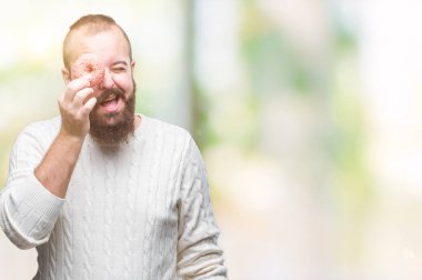 Genç beyaz hipster adam ayakta ve diş gösterilen kendine güvenen bir gülümsemeyle gülümseyerek mutlu bir yüz ile izole arka plan üzerinde tatlı çörek yeme
