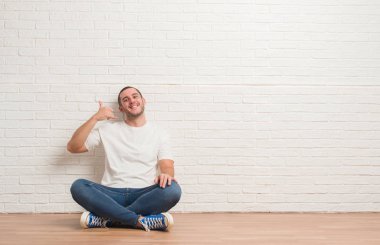 Beyaz tuğla duvar gülümseyen üzerinde katta oturan genç beyaz adam el ve parmak telefonda konuşmak ile telefon jest yapıyor. Kavramları iletişim.