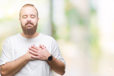 Üzerinde gülümseyen izole arka plan ile rahat t-shirt giyen genç beyaz hipster adam kapalı gözler ve yüzünde minnettar jest ile göğüs üzerinde eller. Sağlık kavramı.