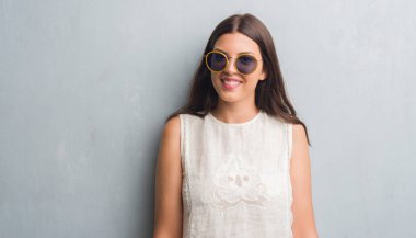 Genç esmer kadın ayakta ve diş gösterilen kendine güvenen bir gülümsemeyle gülümseyerek mutlu bir yüz ile retro güneş gözlüğü takıyor grunge gri duvarın üzerinden