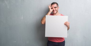 Mutlu yüz el parmaklarının arasından seyir göz üzerinde ok işaretiyle yapıyor gülümseyen ile boş bir afiş tutarak gri grunge duvarın üzerinden beyaz delikanlı