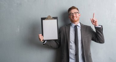 Genç redhead iş adamı gri grunge üzerinde tutarak Pano bir fikirle sürpriz duvar veya gülen yüz, bir numara ile işaret parmağı soru