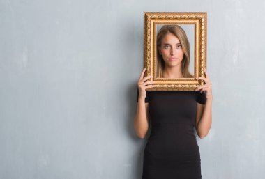 Beautiful young woman over grunge grey wall holding vintage frame with a confident expression on smart face thinking serious clipart