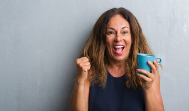 Duygu tezahürat gurur çığlık ve zafer ve başarı çok heyecanlı, kutluyor kahve içme gri grunge duvardan ayakta orta yaş İspanyol kadın