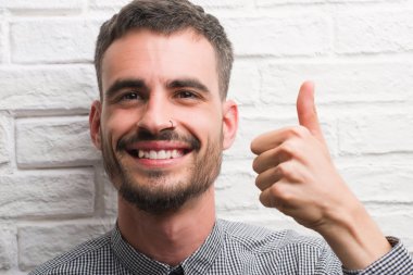 Genç yetişkin adam beyaz tuğla duvardan mutlu büyük gülümseme ok işareti, başparmak ile parmak, mükemmel işareti yapıyor ayakta