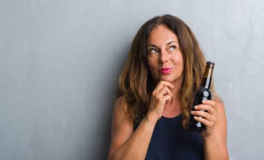 Bira şişe ciddi bir yüz düşünme hakkında soru, çok karışık fikir tutan gri grunge duvarın üzerinden duran orta yaş İspanyol kadın