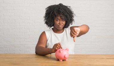 Kızgın yüz, eksi işareti aşağı antipati başparmak ile gösterilen, ret kavramı ile piggy banka holding masada oturan genç Afro-Amerikan kadın
