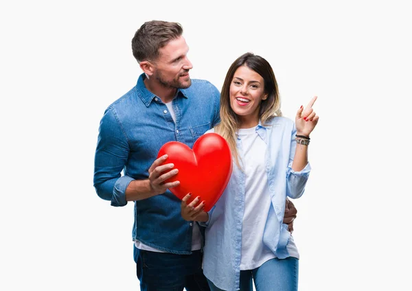 年轻夫妇在爱持有红色心脏在孤立的背景非常高兴地指着手和手指到一边 — 图库照片