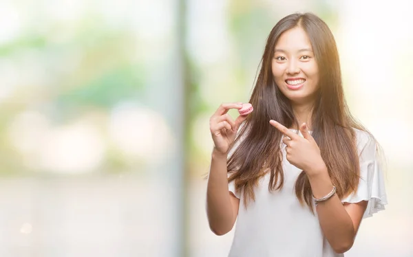 非常に幸せな手と指で指している孤立した背景の上甘いピンクのマカロンを食べる若いアジア女性 — ストック写真