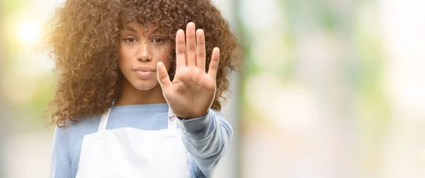 African american shop owner woman wearing an apron annoyed with bad attitude making stop sign with hand, saying no, expressing security, defense or restriction, maybe pushing