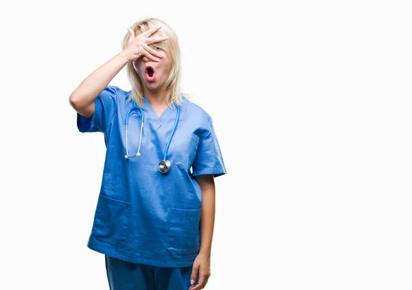 年轻美丽的金发医生妇女穿着医疗制服在孤立的背景下偷看在休克覆盖面和眼睛用手 看着通过手指尴尬的表情 — 图库照片