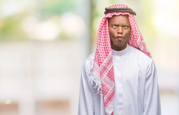 Young arabic african man wearing traditional keffiyeh over isolated background making fish face with lips, crazy and comical gesture. Funny expression.