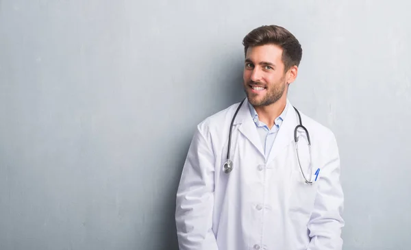 Handsome young doctor man over grey grunge wall looking away to side with smile on face, natural expression. Laughing confident.