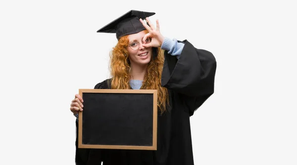 身に着けている赤毛の若い学生女性卒業制服持株黒板指を通して見る目に手で サインをして笑っている幸せそうな顔 — ストック写真