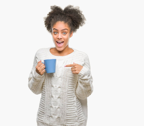 Молодая афро-американка, высунувшая чашку кофе на изолированном фоне, очень довольна, указывая рукой и пальцем.
