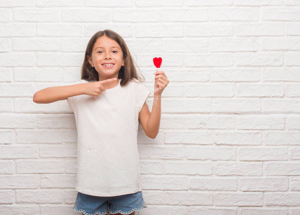 Young hispanic kid over white brick wall eating red heart lollipop candy very happy pointing with hand and finger
