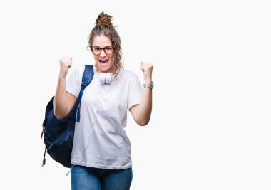 Sırt çantası ve kulaklık üzerinde giyen genç esmer öğrenci kız şaşırmış ve hayret için başarı ile silah kaldırdı kutluyor arka plan izole ve gözleri açık. Kazanan kavramı.