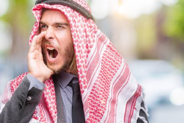 Genç yakışıklı Arap adam uzun saçlı bağırarak ve ağzı üzerinde el ile yan için çığlık izole arka plan üzerinde keffiyeh giyiyor. İletişim kavramı.