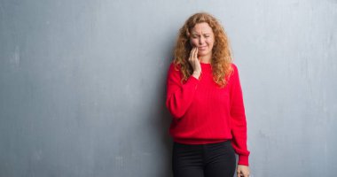 Genç redhead gri grunge duvar giyen üzerinde kırmızı kadın kazak diş ağrısı veya dişlerde dişlerle ilgili hastalık nedeniyle ağız acı ifadesi ile el ile dokunmak. Diş hekimi kavramı.