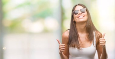 Güneş gözlüğü başarı işareti el ile pozitif jest yapıyor giyen genç güzel İspanyol başparmak gülümseyen ve mutlu. Neşeli bir ifadeyle, kazanan jest kameraya bakıyor.