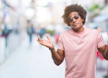Afro Amerikalı adam izole arka plan clueless ve şaşkın bir ifadeyle kollar ve eller kaldırdı üzerinde güneş gözlüğü takıyor. Şüphe kavramı.