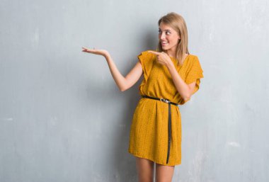 Bir elbiseyle hayrete ve el ile sunulması ve parmak ile işaret ederken kameraya gülümseyen grunge gri duvar üzerinde duran güzel genç kadın.