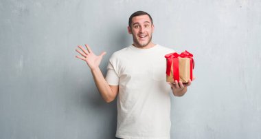 Çok mutlu ve heyecanlı, hediye tutarak gri grunge duvar büyük gülümseme ile çığlık ve ellerini kaldırdı kazanan ifade kutluyor zafer üzerinde beyaz delikanlı