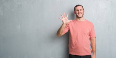 Beyaz delikanlı gösterilen ve parmakları ile işaret gri grunge duvarın üzerinden beş kendinden emin ve mutlu gülümseyerek iken numara.