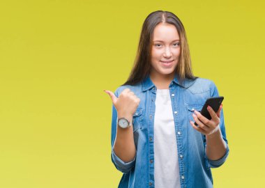 Genç güzel beyaz kadın manifatura işaret ve başparmak kadar mutlu yüz gülümseyen tarafı ile gösteren izole arka plan üzerinde Smartphone kullanarak ileti gönderme