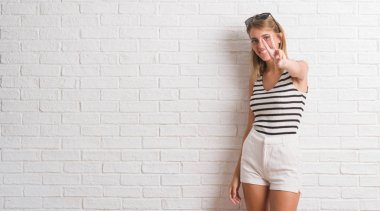 Genç güzel sarışın kadın parmaklarını zafer işareti yaparken gösterilen kamera seyir gülümseyen beyaz tuğla duvarın üzerinden. İki numaralı.