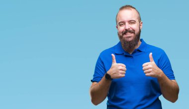 El ile pozitif jest yapıyor izole arka plan başarı işareti üzerinde mavi gömlek giymiş genç beyaz hipster adam başparmak gülümseyen ve mutlu. Neşeli bir ifadeyle, kazanan jest kameraya bakıyor.