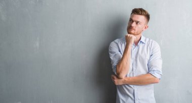 Genç redhead iş adamı çene düşünme hakkında soru, dalgın ifade üzerine el ile gri grunge duvarın üzerinden. Düşünceli yüzüyle gülümsüyor. Şüphe kavramı.