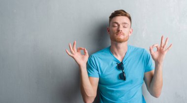 Genç Kızıl saçlı adam gündelik kıyafeti giymiş gri grunge duvarın üzerinden rahatla ve gülümseyen gözlerle parmak ile meditasyon jest yapıyor kapalı. Yoga kavramı.
